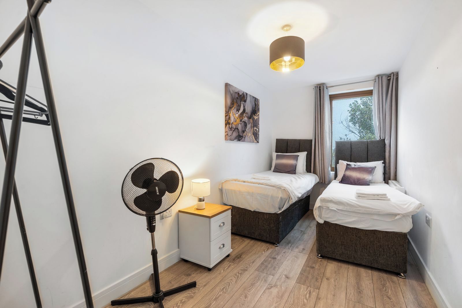 Kompakti hotellihuone, jossa on kaksi sänkyä, lattiatuuletin, yöpöytä lampulla ja ikkuna, jossa on verhot.