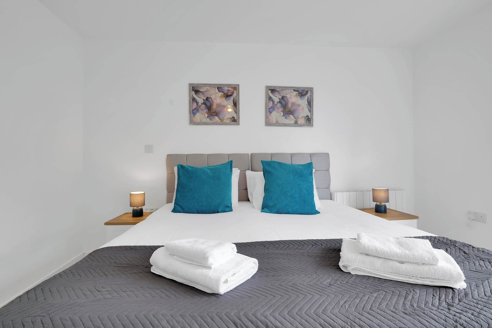Ett hotellrum med en säng, två kuddar, två sänglampor, en filt och två tavlor på väggen.