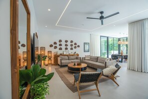 Ein modernes Wohnzimmer mit Sofa, Couchtisch und Sesseln, ein Essbereich mit Tisch und Stühlen sowie ein großes Fenster mit Blick auf Grünflächen.