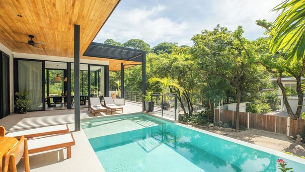 Un área de piscina moderna al aire libre con sillones de descanso, una pérgola de madera y vistas a exuberante vegetación.