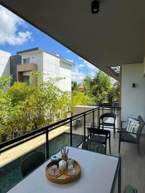 Un balcón con mesa para dos, baranda de vidrio y vista a edificios vecinos y follaje.