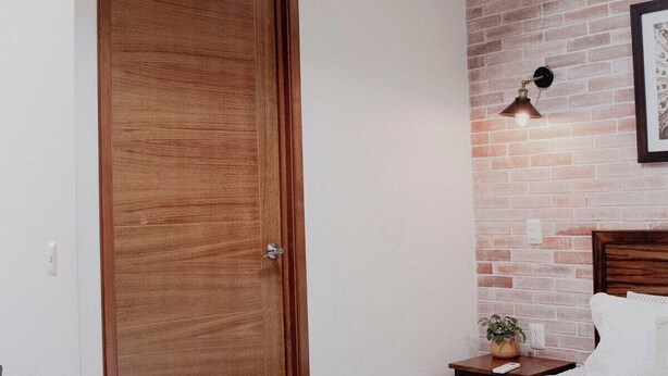 Een slaapkamer met een houten deur, een bakstenen muur, een nachtkastje met een lamp en een bed met een witte dekbedovertrek.