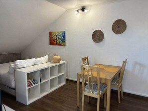 Ein Schlafzimmer mit Bett, einem weißen Bücherregal, einem Esstisch mit Stühlen und einer Deckenleuchte.