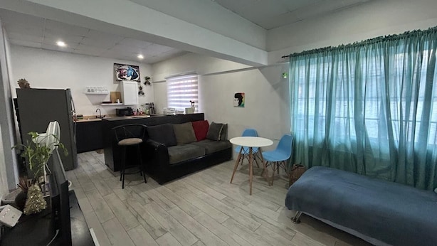 A compact living space with a kitchenette, a sofa, a dining table with chairs, and a large window with curtains. - The Cosy Konak (Port of Spain)