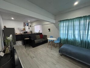 Un espace de vie compact comprenant un coin cuisine, un canapé, une table à manger avec des chaises et une grande fenêtre avec des rideaux.