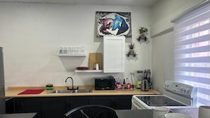 A compact kitchen with a microwave, stove, and sink. There is a dining table with chairs and a coffee table. A painting hangs on the wall above the sink. - The Cosy Konak (Port of Spain)