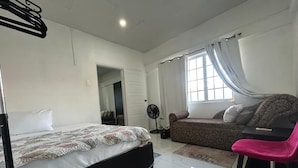 A bedroom with a bed, a sofa, a chair, a fan, and a window with curtains. - The Cosy Konak (Port of Spain)