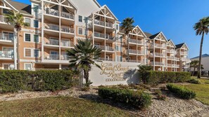 A multi-story condominium building with balconies, palm trees, and a sign reading "The Grand Beach Resort Condominium."