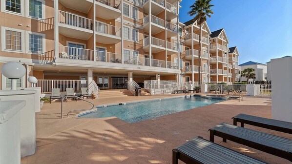 A multi-story hotel with a pool area, outdoor seating, and a palm tree.