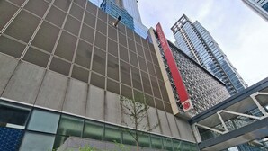 Ein modernes Gebäude mit einer gitterartigen Fassade, einer auffälligen roten vertikalen Beschilderung und einem Baum davor.