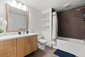 Ein modernes Badezimmer mit einem Holzwaschtisch, einer weißen Toilette, einer Badewanne mit Duschvorhang und einem Spiegel über dem Waschtisch.