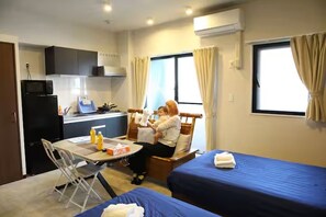 Ein kompaktes Hotelzimmer mit Küchenzeile, zwei Einzelbetten, einem Esstisch und einem Fenster mit Vorhängen.