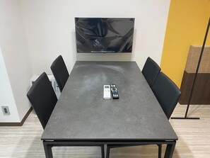 Kokoushuone, jossa on harmaa pöytä, mustat tuolit, seinälle asennettu televisio ja pöydällä kaukosäädin.
