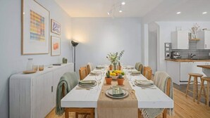 Un coin repas avec une longue table dressée pour six personnes, bordé de meubles blancs et avec une cuisine en arrière-plan.