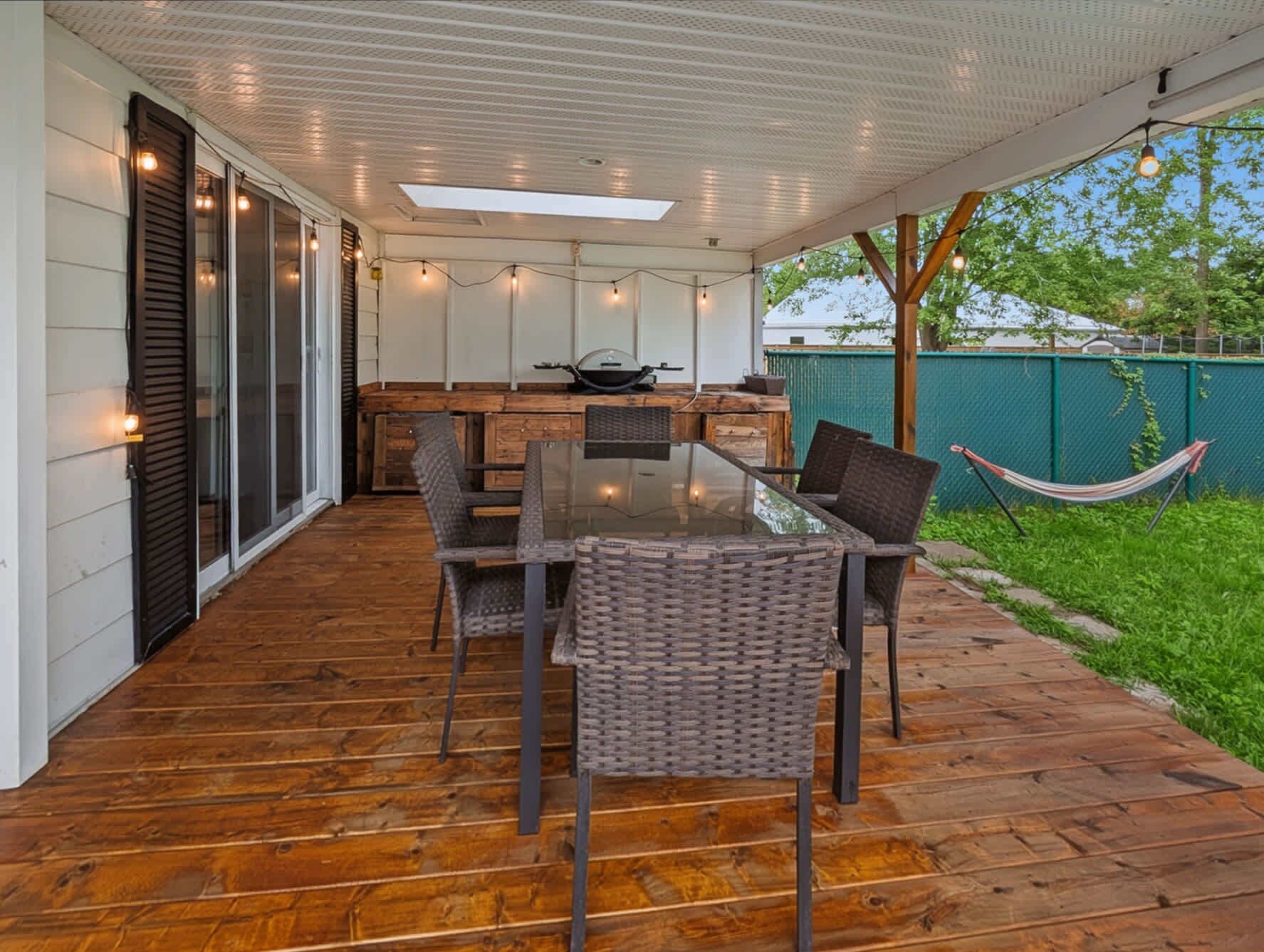 An outdoor patio with a wooden deck, a glass-top table, wicker chairs, and string lights.