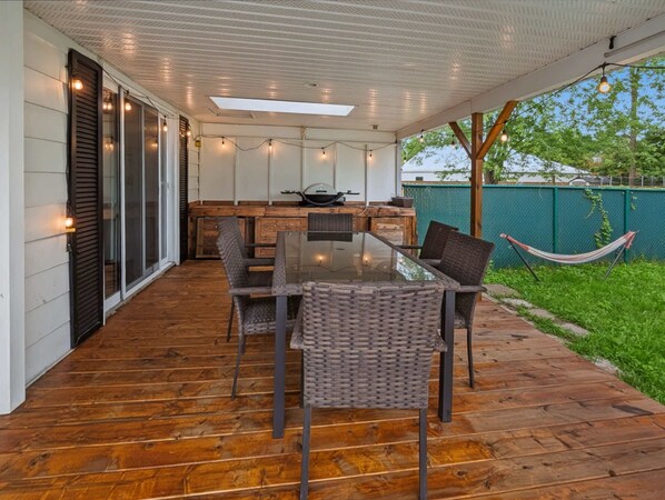 An outdoor patio with a wooden deck, a glass-top table, wicker chairs, and string lights.