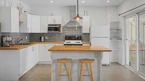 Une cuisine moderne avec des armoires blanches, un plan de travail en bois, un îlot central doté de tabourets, une hotte de cuisine en acier inoxydable et un luminaire suspendu.