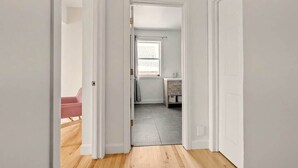Un couloir doté d’un plancher en bois, de murs blancs et d’une porte donnant sur une salle de bain avec une fenêtre.