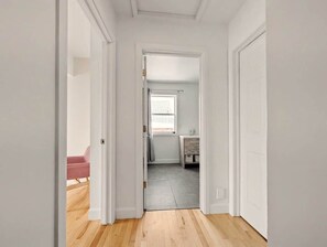 A hallway with a wooden floor, white walls, and a door leading to a bathroom with a window.