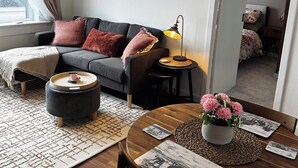 A modern living room with a sofa, coffee table, and a dining table with a vase of flowers. There are framed pictures on the wall and a window with curtains.