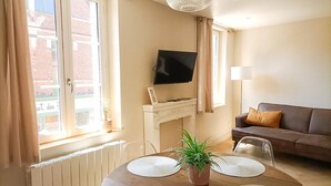 A living room with a fireplace, a television mounted above it, a sofa, and a dining table with placemats. - Roses, Veules les roses close to the beaches (Veules-les-Roses)