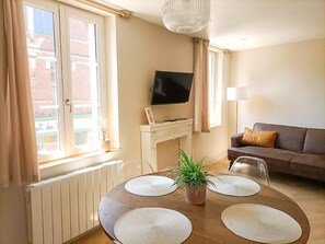 A living room with a fireplace, a television mounted above it, a sofa, and a dining table with placemats. - Roses, Veules les roses close to the beaches (Veules-les-Roses)