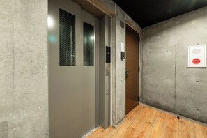 Um corredor com piso de madeira, uma porta cinza com janela e um interfone fixado na parede.