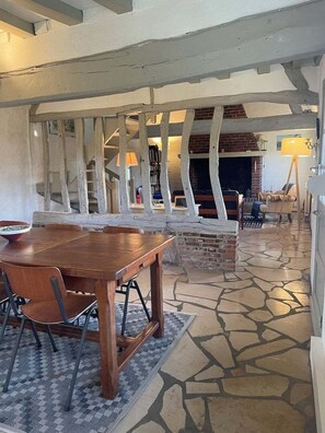 Een ruime woonkamer met een houten eettafel en stoelen, een open haard en een vloer van stenen tegels.