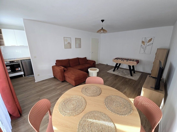 Una sala moderna con un sofá marrón, una mesa redonda de madera con individuales tejidos y un comedor con sillas rosas.