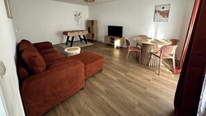 Uma sala de estar com um sofá marrom, uma mesa de centro de madeira, uma televisão e um espaço de refeições com uma mesa redonda e cadeiras.