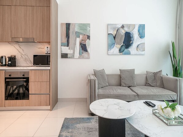 A modern kitchen with a built-in oven and microwave, a grey sofa, abstract art on the wall, and a round coffee table with a vase of flowers. - Beachfront Living With Marina Skyline Views (Dubai)