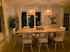 A dining area with a wooden table, white chairs, two wicker pendant lights, a potted plant, and a deer head decoration on the wall.