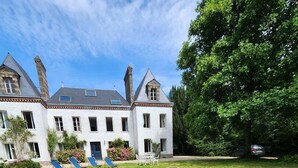 Un grande edificio bianco con numerosi camini e finestre, circondato da un prato curato con mobili da giardino.