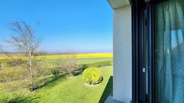 Un balcon donnant sur une pelouse verdoyante, un champ jaune et un ciel bleu dégagé.