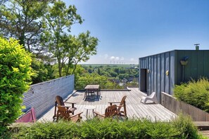 A wooden deck with outdoor furniture, a view of wind turbines, and a green building in the background. - La villa California - Veules-les-Roses (Veules-les-Roses)