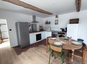 Une cuisine avec des meubles blancs, un réfrigérateur gris, une table à manger en bois avec des chaises et un luminaire suspendu.