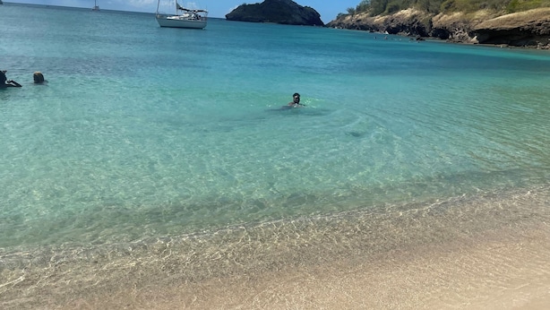 Una playa con agua turquesa cristalina, una orilla arenosa y un velero a lo lejos.