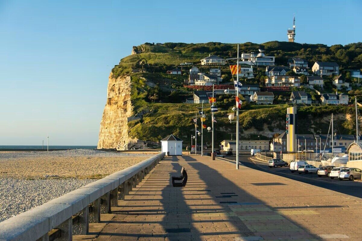Una strada costiera con un molo in pietra che conduce a una spiaggia, una collina con delle case e un faro sullo sfondo.