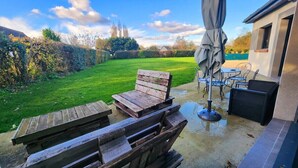 An outdoor patio area with wooden benches and a table, a covered umbrella, and a view of a green lawn and trees. - The countryside at the edge of the sea (Manneville-ès-Plains)