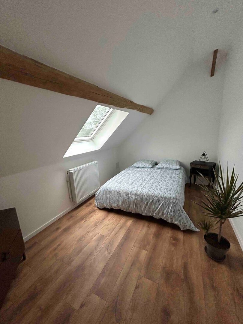 Une chambre avec un plafond en pente, un puits de lumière, un radiateur, un lit, une petite table et une plante en pot.
