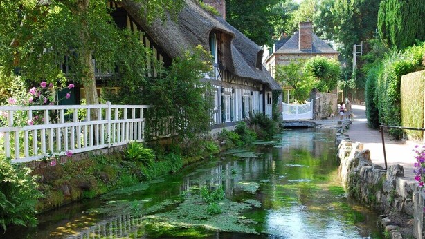 A narrow canal with buildings on one side, a white fence, and a stone wall on the other. - L'Écho de la Nature | Norman Farm • 1ha • Near Veules & Étretat (Sasseville)