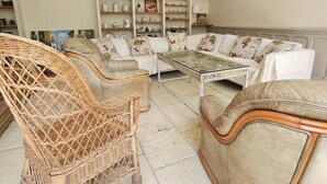A living room with wicker chairs, a glass coffee table, and a sofa with floral cushions. There are shelves with various decorative items and a painting on the wall.