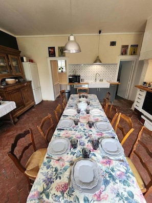 A dining area with a long table set for six, surrounded by wooden chairs. There is a kitchen area with a refrigerator, microwave, and sink in the background.