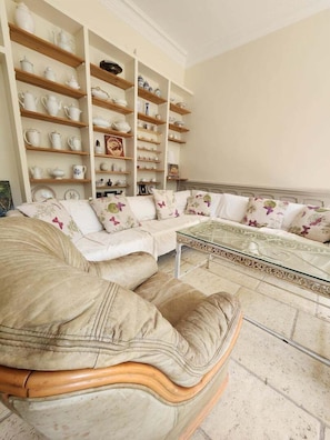 A living room with a white sofa, a glass coffee table, and shelves with white and patterned dishes.
