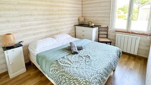 Ein Schlafzimmer mit Holzbalkendecke, einem Bett mit gemusterten Betttextilien, einem Nachttisch, einem Sessel und einem Fenster mit Blick ins Grüne.