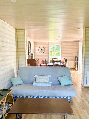 Ein gemütliches Blockhaus-Interieur mit einer Couch, einem kleinen Tisch, einer Essecke mit Tisch und Stühlen sowie einem Fenster mit Grünpflanzen draußen.