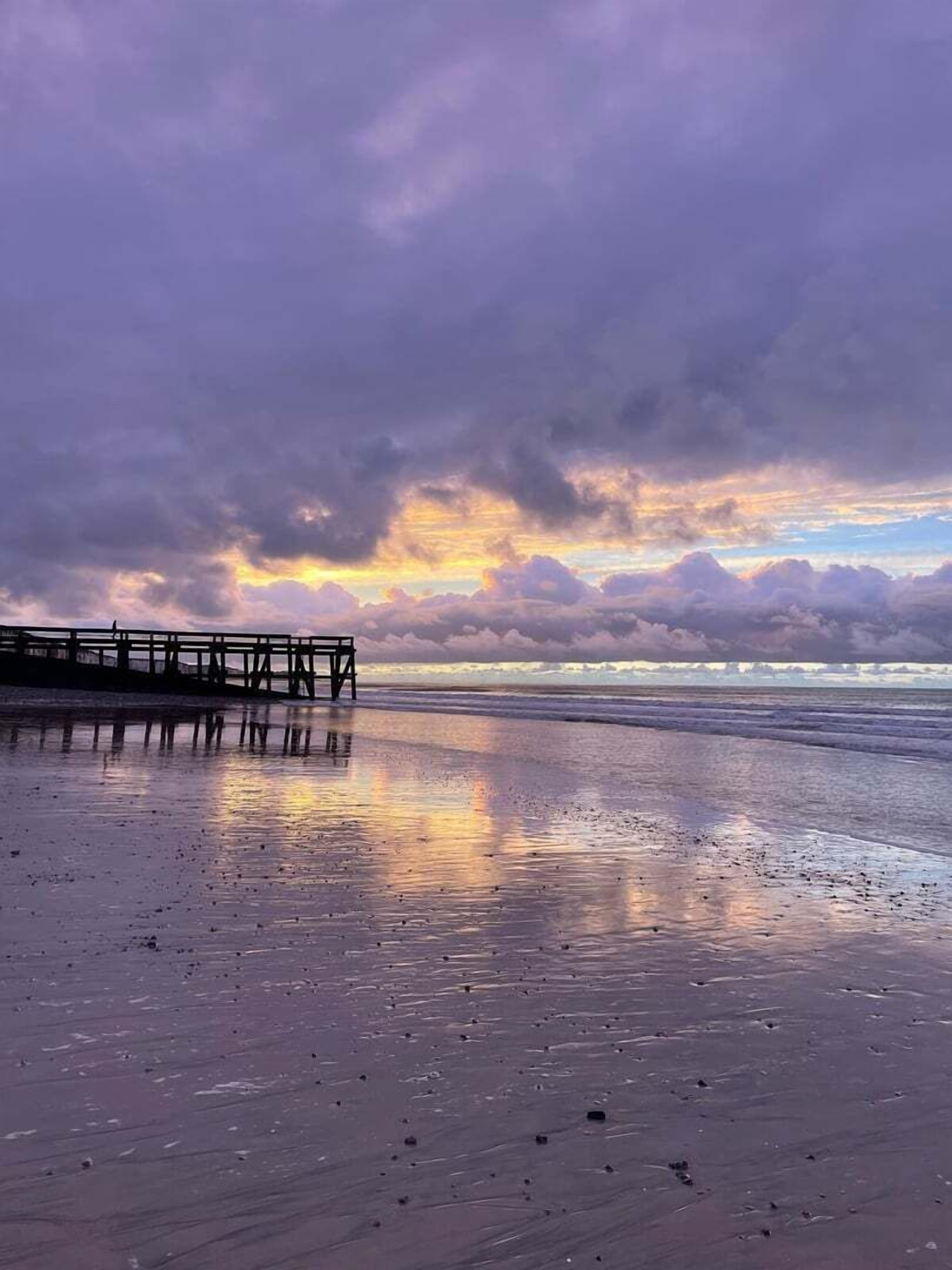 Una playa con un muelle de madera que se adentra en el agua, un mar tranquilo y un cielo al atardecer con nubes.