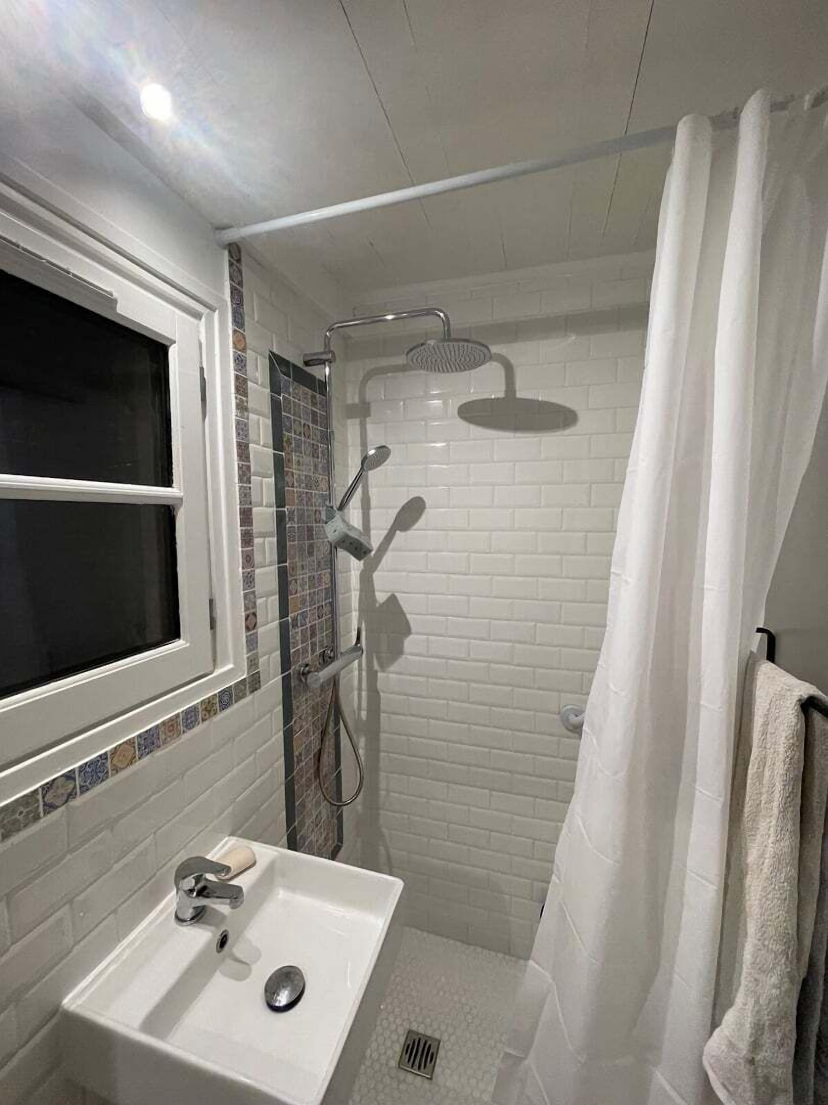 Un baño con lavamanos blanco, ducha con cabezal de ducha de mano y ventana con cortina negra.
