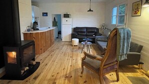 A cozy living room with a fireplace, wooden flooring, a sofa, a chair, and a kitchen area in the background. - Anchoring, cottage near the sea (Sotteville-sur-Mer)