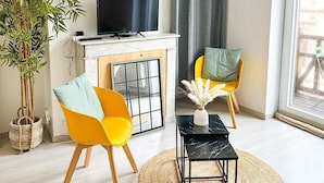 Un salon avec une cheminée, un téléviseur, deux fauteuils, une petite table et une grande fenêtre avec des rideaux.